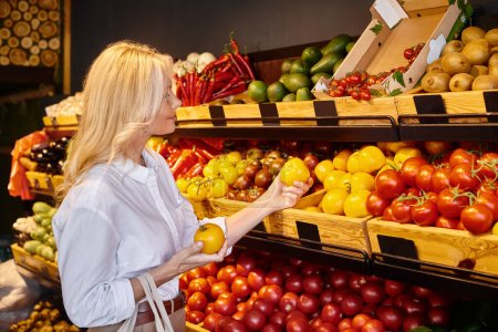 Foto de Mujer madura de buen aspecto en ropa casual recogiendo verduras frescas y vibrantes en la tienda de comestibles - Imagen libre de derechos