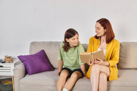 smiling woman reading book about sex education to teenage daughter on cozy couch in living room