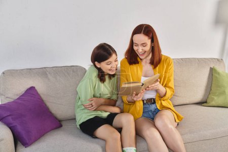 smiling woman reading book about sex education to teenage daughter on cozy couch in living room