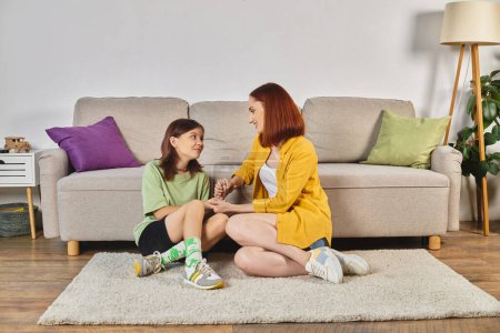 femme souriante avec fille adolescente tenant la main et parlant d'éducation sexuelle sur le sol à la maison