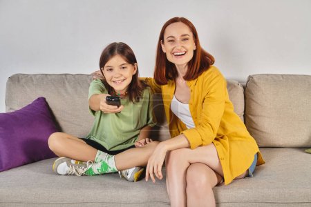 Téléchargez les photos : Femme riante avec fille adolescente regardant un film sur un canapé confortable dans le salon moderne, loisirs - en image libre de droit