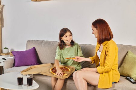 smiling woman giving piece of pizza to teenage daughter near glasses with soda in living room