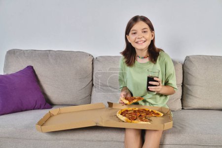 joyful teenage girl looking at camera while sitting on couch with pizza and soda, meal time