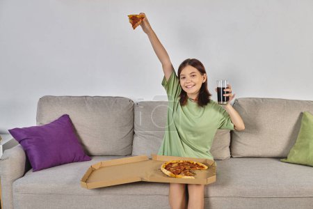 adolescente emocionada sentada en el sofá con pizza y un vaso de cola y mirando a la cámara, hora de la comida