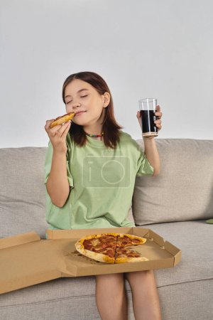 delighted teenage girl with closed eyes holding pizza and glass of soda on couch at home, enjoyment