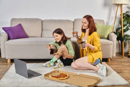 happy mother and teen daughter with pizza and soda watching movie on laptop on floor in living room