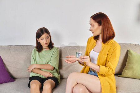 Téléchargez les photos : Mère montrant un préservatif à une adolescente confuse lors d'une conversation à la maison, éducation sexuelle - en image libre de droit