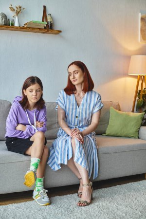 thoughtful woman looking at frustrated teenage daughter on couch in living room, care and support