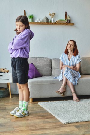 offended teen girl standing with folded arms near displeased mother on couch, family conflict