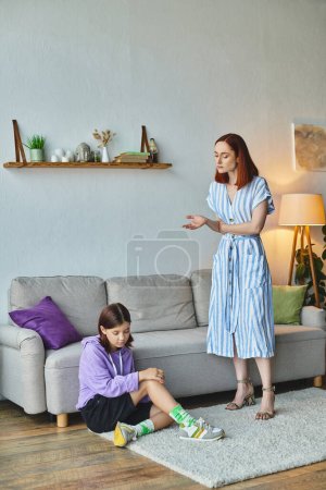 femme sérieuse parlant à une adolescente frustrée assise sur le sol dans le salon, écart de génération