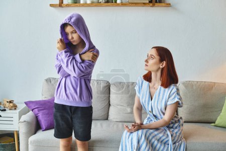 worried woman talking to offended daughter standing in hood in living room, generation gap