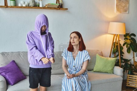 worried and upset woman talking to offended daughter hiding in hood in living room, generation gap