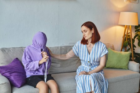 caring mother calming depressed teenage daughter obscuring face with hood on couch at home, support