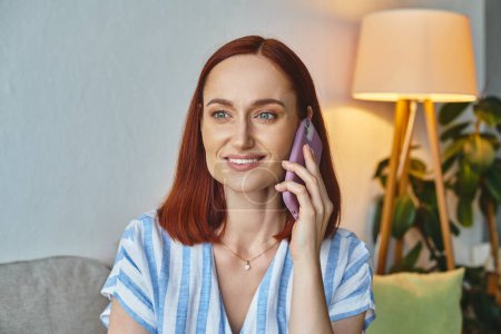 happy stylish woman smiling and talking on smartphone in modern living room, domestic scene