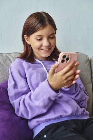 Téléchargez les photos : Joyeuse adolescente naviguant sur les médias sociaux tout en étant assis sur un canapé confortable dans le salon, loisirs - en image libre de droit