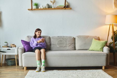 Téléchargez les photos : Joyeuse adolescente naviguant sur les médias sociaux tout en étant assis sur un canapé confortable dans le salon moderne - en image libre de droit