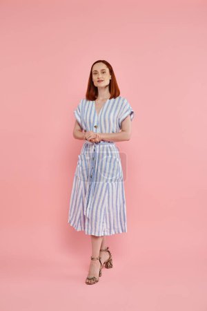full length of stylish redhead woman in striped dress standing and looking at camera on pink