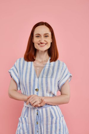 cheerful redhead woman in striped dress looking at camera on pink backdrop, studio portrait