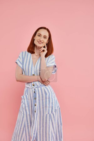 cheerful redhead woman in striped dress looking at camera on pink backdrop, studio portrait