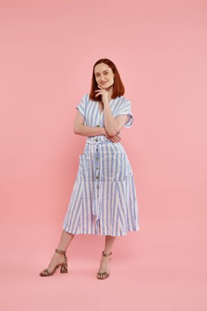 full length of smiling redhead woman in striped dress looking at camera on pink backdrop, fashion