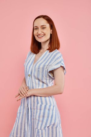 joyful and trendy redhead woman in striped dress looking at camera on pink backdrop, studio portrait