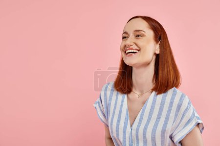 excited redhead woman in striped dress looking away and laughing on pink backdrop, happiness