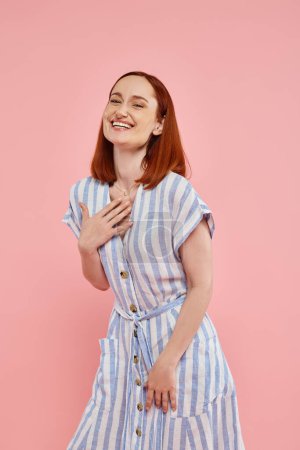 excited redhead woman in trendy striped dress looking at camera and laughing on pink backdrop
