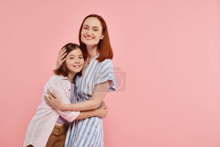 mujer feliz con hija adolescente en elegante atuendo casual mirando a la cámara en el telón de fondo rosa