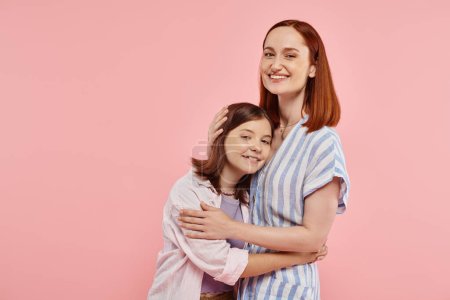 mujer feliz con hija adolescente en elegante atuendo casual mirando a la cámara en el telón de fondo rosa