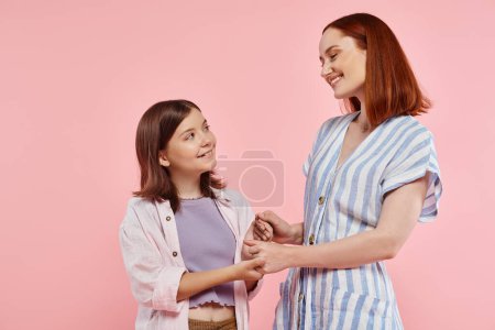 mujer alegre y de moda con hija adolescente cogida de la mano y sonriente el uno al otro en rosa