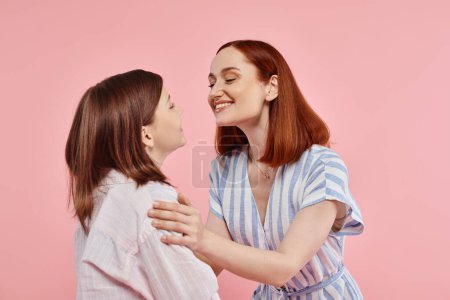 joyful and stylish woman with teenage daughter smiling at each other on pink backdrop in studio