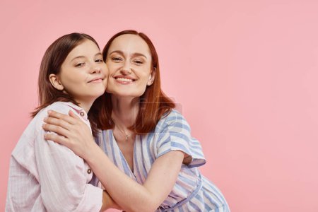 mujer alegre con hija adolescente en elegante atuendo casual abrazando y mirando a la cámara en rosa