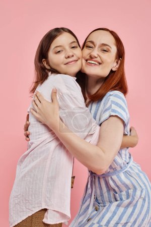 joyful woman with teenage daughter in stylish casual attire embracing and looking at camera on pink