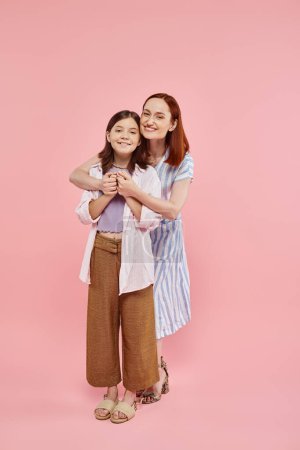 mujer alegre abrazando hija adolescente y sosteniendo sus manos en el telón de fondo rosa, longitud completa