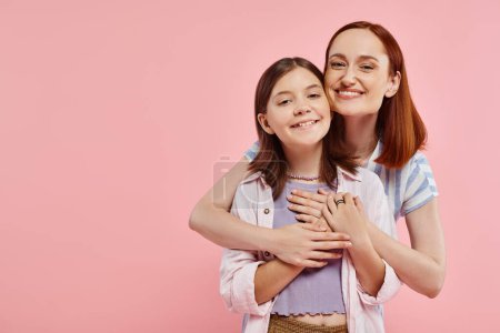 cheerful and stylish woman embracing happy teenage daughter and looking at camera on pink backdrop