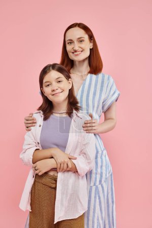 fröhliche und stilvolle Frau umarmt Schultern ihrer Teenager-Tochter und schaut in die Kamera auf rosa