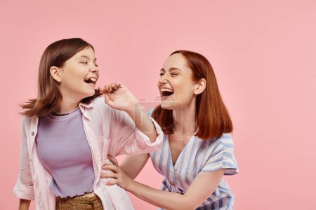 excited stylish woman with cheerful teenage daughter having fun and laughing on pink backdrop