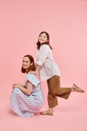 Téléchargez les photos : Femme élégante gaie assise sur des hanches près de fille adolescente posant sur fond rose, amusant et joie - en image libre de droit