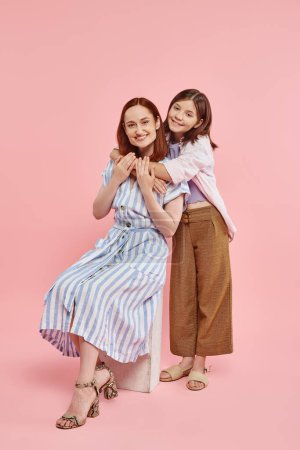 happy teenage girl embracing stylish mother sitting on stone block on pink backdrop, love and unity