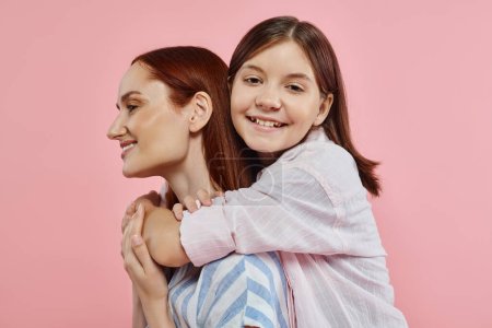 cheerful teenage girl embracing stylish mother smiling on pink backdrop, unity of mom and daughter