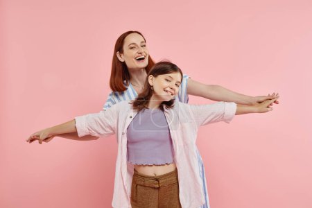 fröhliche und stilvolle Frau hält Hand in Hand mit frühgeborener Tochter auf rosa Hintergrund, glückliche Familie