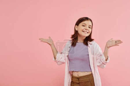 happy teenage girl in stylish casual attire posing with open arms and looking at camera on pink