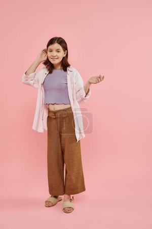joyful and stylish teenage girl pointing with hand and looking at camera on pink, full length