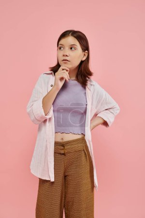 thoughtful and stylish teenage girl standing with hand on hip and looking away on pink backdrop
