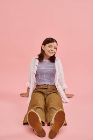 full length of joyful teenage girl in trendy casual attire sitting and smiling at camera on pink