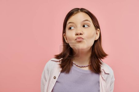 positive and curious teenage girl in trendy clothes pouting lips and looking away on pink backdrop