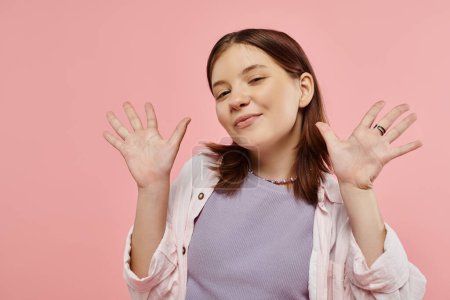cheerful teenage girl in stylish casual clothes waving hands and looking  at camera on pink backdrop