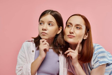 thoughtful woman with teenage girl in stylish casual clothes looking away on pink backdrop