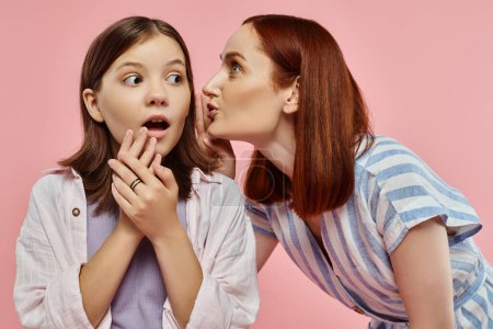 excited mother telling secret to amazed teenage daughter with open month on pink backdrop, gossip