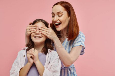 joyful and stylish woman covering eyes of preteen daughter while playing guess who game on pink